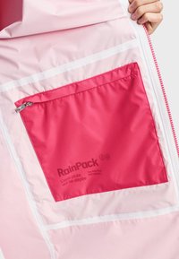 Main tenant une veste rose clair ouverte, révélant une poche intérieure zippée rose vif étiquetée « RainPack » avec texte en français et en anglais.
