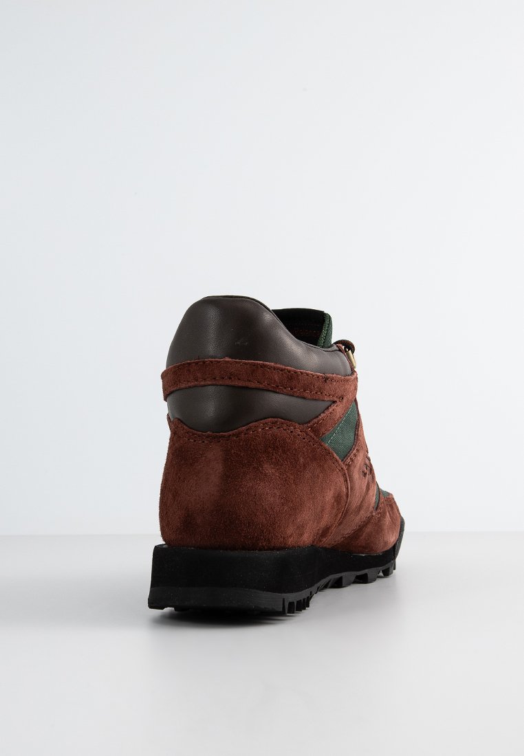 Chaussure de randonnée en daim marron avec des accents en tissu vert, semelle en caoutchouc et support de cheville rembourré, comportant une boucle en métal pour le laçage.