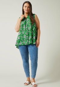 Blusa verde senza maniche con un motivo floreale bianco, design drappeggiato, orlo a contrasto. Abbinate a jeans skinny blu chiaro e sandali.
