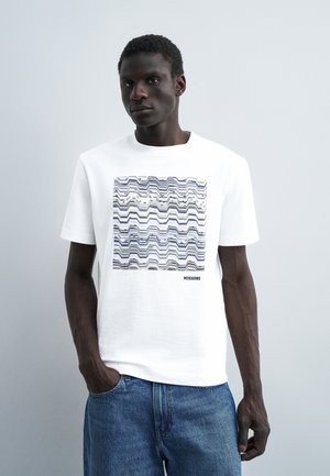 Mężczyzna w białej koszulce z krótkim rękawem z wielokolorowym wzorem zygzakowatym w kwadraty i napisem "MISSONI", połączonej z niebieskimi dżinsami.