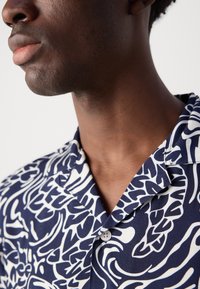 Camisa de manga curta azul-marinho com um padrão abstrato branco, gola camp e um detalhe de botão. Confeccionada em tecido leve.