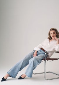 Camisa blanca de botones, jeans de denim anchos azules, tacones negros de Punta, cinturón marrón, sentado en una silla de chrome minimalista, fondo neutro.