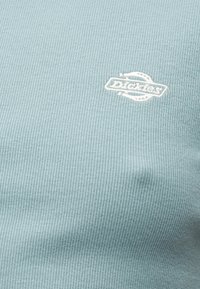 Ανοιχτό μπλε ριμπ ύφασμα με μικρό λευκό κεντημένο λογότυπο "Dickies" στην περιοχή του στήθους ενός ενδύματος.