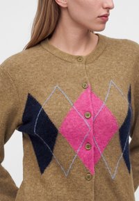 Femme portant un cardigan marron avec des motifs losanges bleu marine et rose et des boutons beiges, visible des épaules jusqu'au milieu du torse.