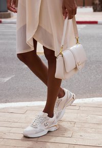 Vita sneakers med guldaccenter, med en texturerad ovandel, rund tå och tjock sula. En liten logotyp syns på sidan.