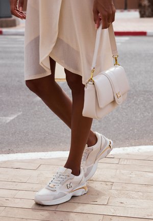 Witte sneakers met gouden accenten, voorzien van een gestructureerde bovenkant, ronde neus en dikke zool. Een klein logo is zichtbaar aan de zijkant.