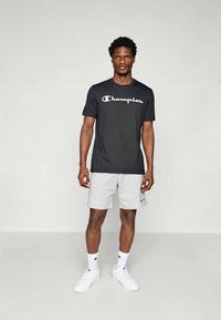 Schwarzes Kurzarm-T-Shirt mit weißem "Champion"-Logo, kombiniert mit grauen Shorts. Das Outfit umfasst weiße Sportschuhe und Socken.