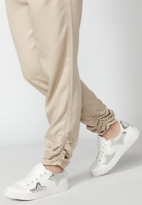 Pantalon beige avec poignets froncés associé à des baskets blanches ornées de motifs d’étoiles argentées, porté sur un fond clair.