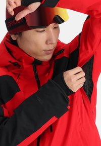 Rote und schwarze Skijacke mit Reißverschlüssen, netzartiger Fütterung, verstellbare Kapuze und elastischen Bündchen. Tragen mit verspiegelten Skibrillen, die das schlanke Design unterstreichen.