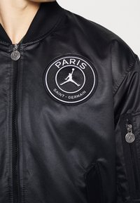 Black bomber jacket with a smooth satin finish, featuring a circular embroidered patch with "PARIS" and "SAINT-GERMAIN" text. Zipper detail on sleeve.