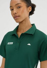 Ciemnozielona koszulka polo z kołnierzykiem, z małym białym logo po lewej stronie klatki piersiowej oraz białym akcentem po boku. Gładka faktura tkaniny.