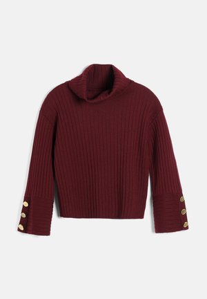Femme aux longs cheveux foncés portant un pull côtelé bordeaux, un pantalon assorti, une ceinture imprimé serpent et des talons, debout les bras croisés.