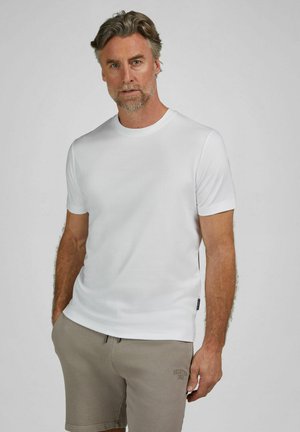 Man met grijs haar en baard, gekleed in een eenvoudig wit T-shirt met korte mouwen en beige korte broek, met gedeeltelijk in de zakken gestoken handen, tegen een neutrale achtergrond.