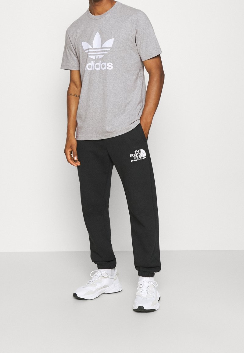 Man som bär en grå Adidas T-shirt, svarta The North Face joggers och vita sneakers, står med en hand i fickan.