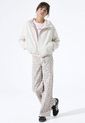 Leichte cremefarbene Jacke mit Kapuze, kombiniert mit locker sitzender Hose im Leopardenmuster in Beige- und Grautönen. Schwarze Turnschuhe runden das Outfit ab.