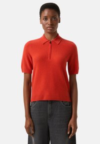 Roter Pullover mit kurzen Ärmeln und Reißverschluss am Ausschnitt, strukturiertem Stoff, geripptem Saum und leichtem Stretch, kombiniert mit dunkelgrauer Jeans.