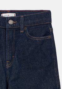 Pantalones vaqueros de mezclilla azul oscuro con cierre de botón, costuras de cobre y una etiqueta con el logo cerca de la cintura, con un diseño clásico de cinco bolsillos.