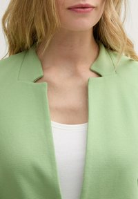 Veste côtelée vert clair avec un col structuré et une ouverture devant, superposée à un haut blanc, mettant en valeur sa texture et son contraste de couleurs.
