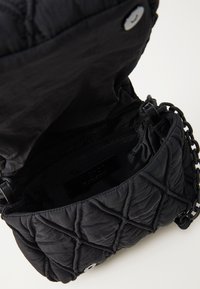 Schwarze gesteppten Handtasche mit Magnetverschluss, geöffnet und zeigt das Innenfach mit Reißverschluss und Kettenriemen-Details auf einer hellen Oberfläche.