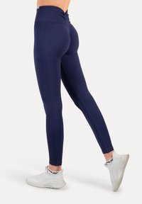Granatowe legginsy z wysokim stanem, z marszczeniem z tyłu. Wykonane z elastycznego materiału, zestawione z białymi butami sportowymi.