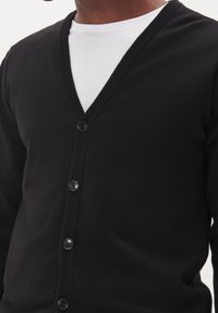 Černý kardigan s véčkovým výstřihem, žebrovaným límcem a šesti předními knoflíky. Nosený přes bílé tričko, které zdůrazňuje hladkou pletenou texturu.