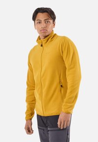 Veste en polaire jaune avec un col montant, une fermeture éclair frontale et une poche latérale zippée. Texture douce et coupe ajustée.