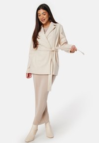 Giacca avvolgente beige con cintura e colletto a revers, abbinata a un vestito beige attillato e stivaletti crema. Texture liscia e vestibilità su misura.