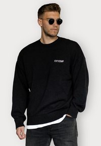Schwarzer übergroßer Strickpullover mit gerippten Bündchen und Saum, mit einem kleinen weißen Logo auf der linken Brust. Trägt man über einem weißen T-Shirt.