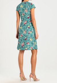 Šaty s květinovým vzorem v teal tkanině, krátkými rukávy a délky do kolen; mají vypasovaný pas a hladkou texturu.