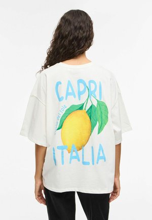 Osoba z długimi kręconymi włosami nosząca białą, oversize koszulkę z dużą cytryną i niebieskim napisem "CAPRI ITALIA" na plecach.