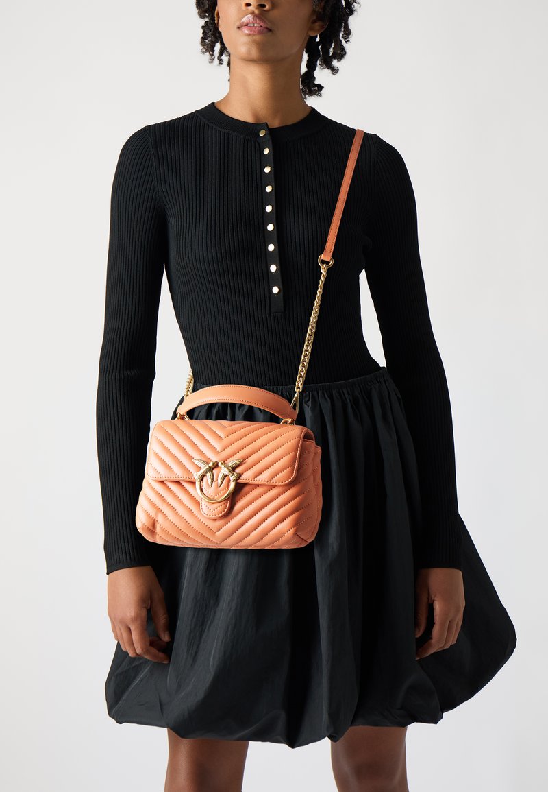 Quiltad orange läder crossbody-väska med ett handtag i guld, kedjesnodd och en strukturerad form, hålls mot en svart klänning.