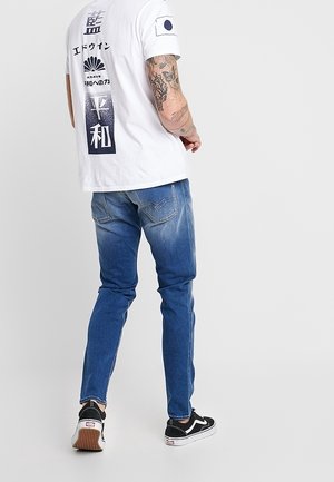 Man met tatoeages die een wit T-shirt met Japanse tekst en symbolen draagt, een blauwe spijkerbroek en zwarte sneakers, staand tegen een witte achtergrond.