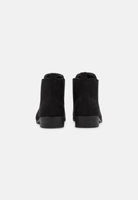 Botas de tobillo de ante negro que presentan un tacón bajo apilado, paneles laterales elásticos y tiradores para facilitar su uso. Textura suave con un diseño minimalista.