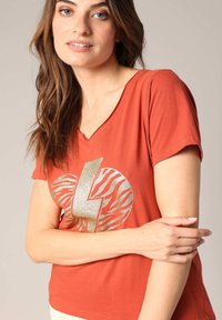 Roest-oranje V-hals T-shirt van zachte stof, met een gouden glitter bliksemschicht en een zebra-print ontwerp aan de voorkant. Korte mouwen.