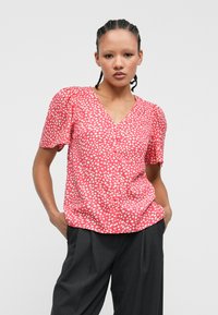 Ροζ κοντομάνικο μπλουζάκι με λευκές βούλες, V-λαιμόκοψη και κουμπιά μπροστά. Συνδυάζεται με σκούρες γκρι παντελόνες, που διαθέτουν άνετη εφαρμογή.