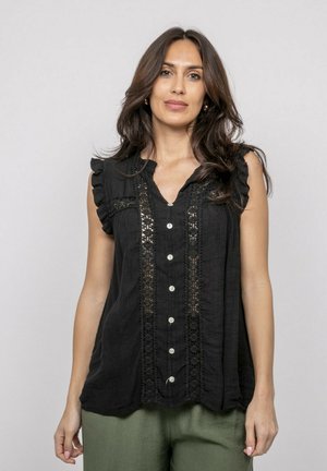 Femme aux longs cheveux noirs portant un chemisier noir sans manches à boutons avec des détails en dentelle et un pantalon vert olive sur un fond uni.