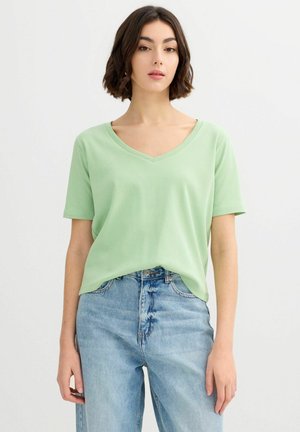 Joven mujer con el pelo corto y oscuro, que lleva una camiseta de cuello en V de color verde claro y vaqueros azules de talle alto, de pie frente a un fondo blanco liso.