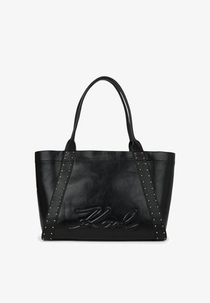 Fekete bőr bevásárlótáska dupla fogantyúkkal, arany tónusú szegecsekkel a pántokon, és a "Karl" aláírás dombornyomott mintájával az elülső panelen.