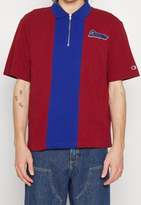 Personne portant une chemise rouge à manches courtes avec une bande verticale bleue et une fermeture éclair à l'avant, associée à un jean bleu, montrant des tatouages sur le bras.