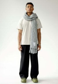 Écharpe en laine grise avec des franges, portée sur un t-shirt beige clair et un pantalon noir plissé, accompagnée de chaussures vertes.