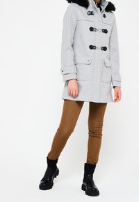 Šedý kapucňový plášť s černým umělým kožešinovým lemem, zapínání na knoflíky a dvěma předními kapsami, v kombinaci s hnědými úzkými kalhotami a černými kotníkovými botami.