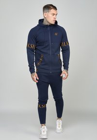 Tuta navy con giacca a zip con cappuccio e pantaloni attillati. Presenta dettagli testuali in oro e un tessuto texture. Sneakers bianche completano il look.