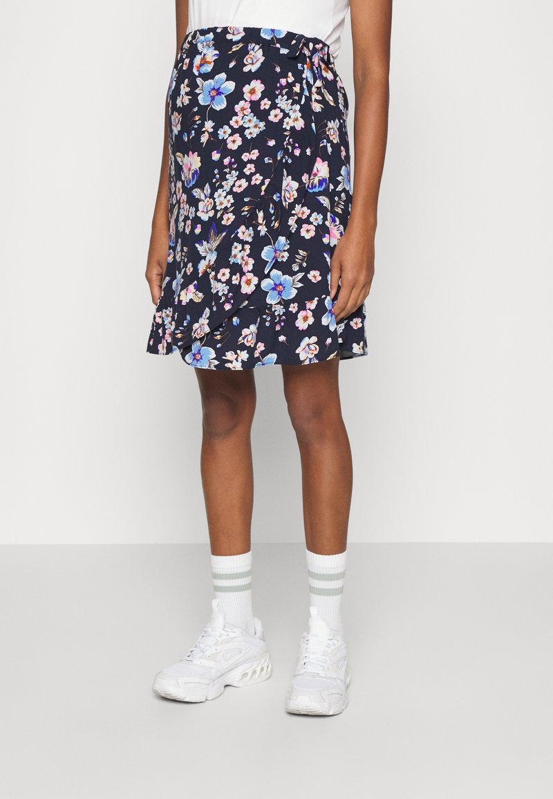 Femme portant une jupe portefeuille bleu marine à motifs floraux, associée à des chaussettes blanches avec deux rayures grises et des chaussures de sport blanches.