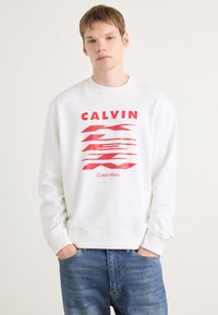 Felpa bianca con un design grafico rosso che presenta il testo "CALVIN" e "Calvin Klein". Ha polsini a costine e uno stile a girocollo.