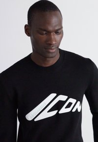 Sort sweater lavet af strikket materiale med et fremtrædende hvidt logo "ICON" på forsiden. Rund halsudskæring og ribbede manchetter. Glat tekstur.