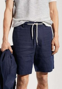 Man wearing navy drawstring shorts and a white striped t-shirt, holding matching navy jacket in left hand.