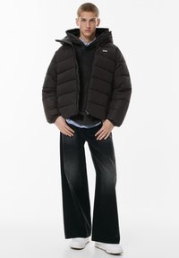 Puffjakke i mørkebrun med horisontalt quiltet design, hætte, lynlås; parret med en mørk sweater, vidde jeans og hvide sko.