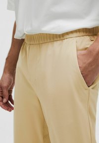 Beżowe spodnie z elastycznym pasem i bocznymi kieszeniami, o gładkiej teksturze i minimalnych detalach szwów. Połączone z białą koszulką.