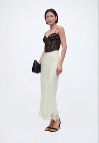 Γυναίκα που φοράει μαύρο κορσέ από δαντέλα, κρεμ σατέν φούστα με δαντελένιο τελείωμα, μαύρα πέδιλα με λουράκια και κρατάει μια μαύρη τσάντα χειρός.