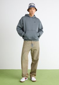 Grauer Kapuzenpullover mit einer Fronttasche und schwarzem Logo, kombiniert mit beigen, lässig geschnittenen Jeans und weißen Sneakers mit roten Akzenten. Blauer karierter Bucket Hat.
